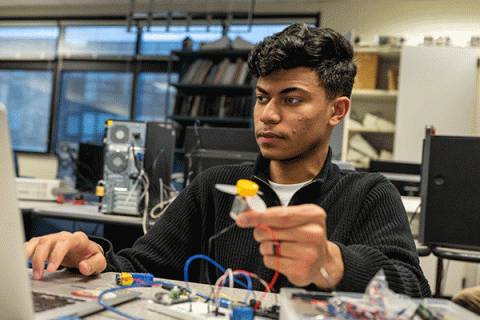 A male student with a dak olive complexion and short black hair sits at a table with a laptop in front of him. He wears a black zipped-up jacket with a white undershirt. Blue and red wiring is visible on the table. He holds a small grey electrical device in his right hand, with a yellow top and grey propellers. Other electrical devices are visible on the table. Computer monitors, a bookcase with stacks of books, and a row of windows are visible in the background. 